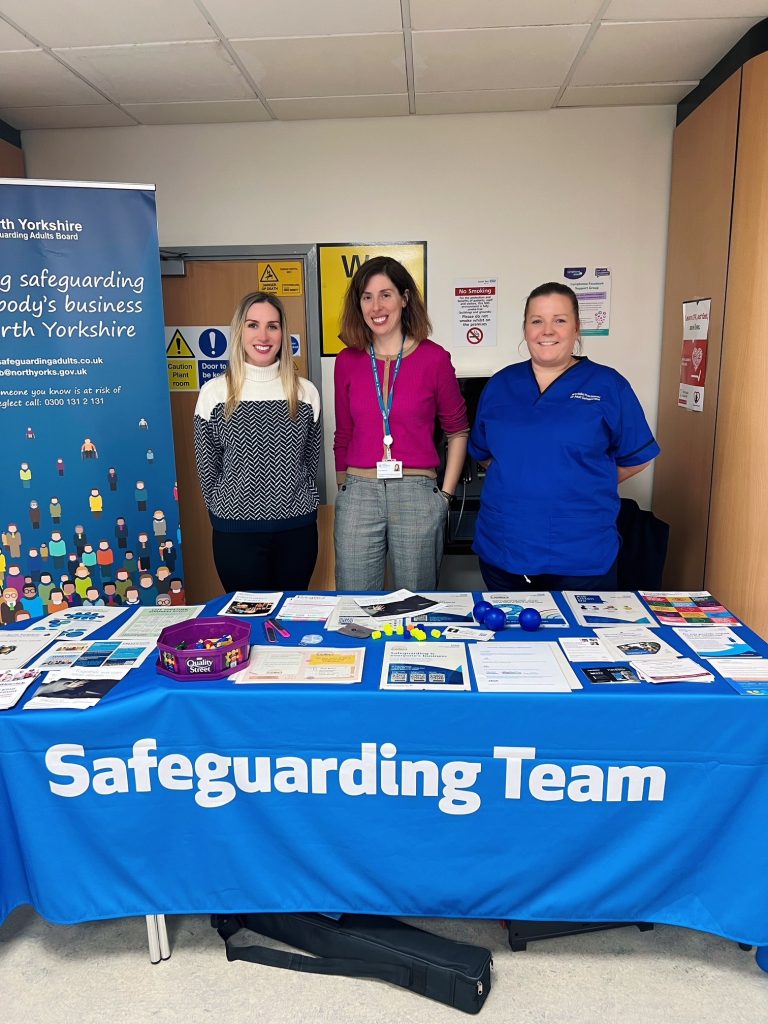 Image of two members of the Safeguarding Adults Board with NHS worker stood behind a table with leaflets and sweets to promote safeguarding adults week. Pop up banner also in the background. 