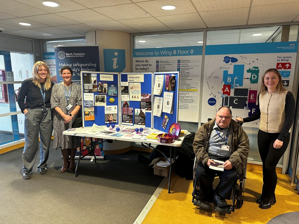Photo of people stood around a table with our Safeguarding Champion. The table has leaflets, sweets and promotional materials such as stress balls to help promote safeguarding adults week. There is also a board with lots of posters on promoting safeguarding. 