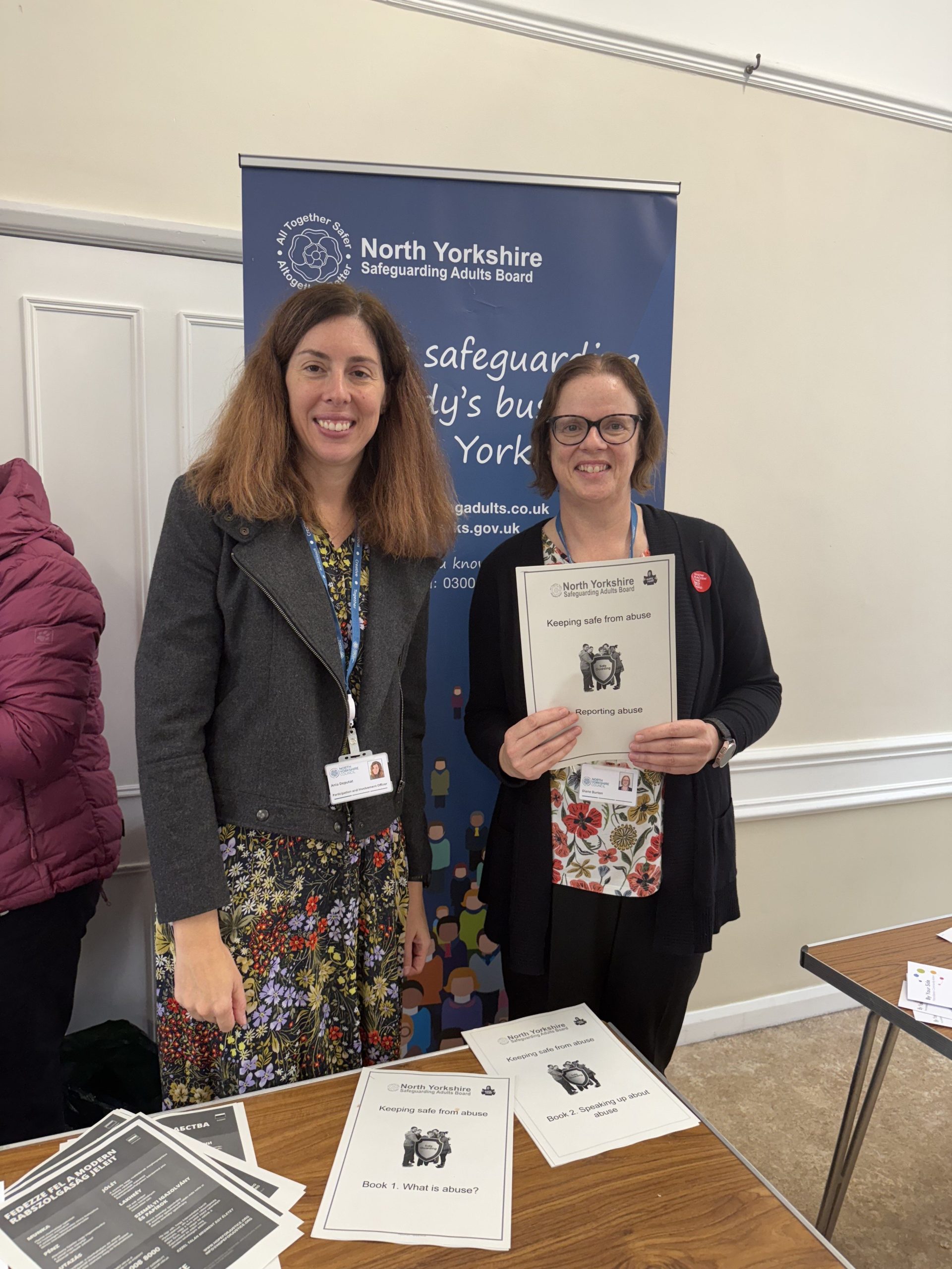 Photo of two people in front of the safeguarding adults pop up banner. One person is holding a piece of paper that says 'keeping safe from abuse' 