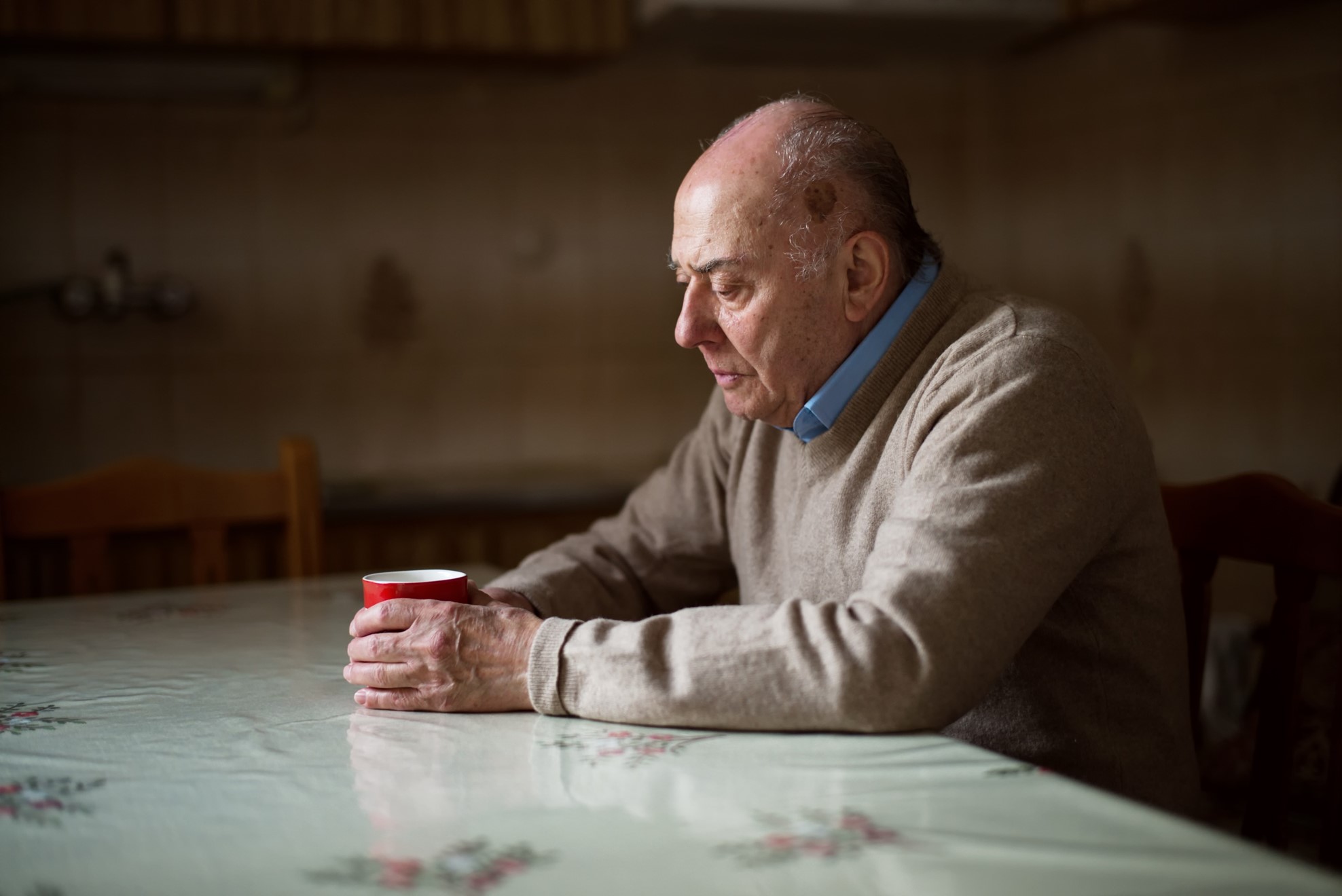 Photo of an elderly man sat at his kitchen table with a sad expression on his face holding a cup of tea