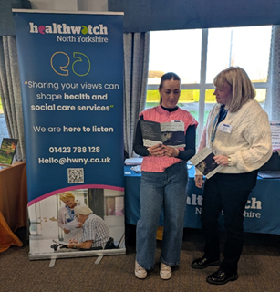 Two women stand side by side next to a vertical pop up banner describing promoting Healthwatch, on organisation who help us to hear peoples opinions. The women are both looking at Safeguarding Adults  Board leaflets. 