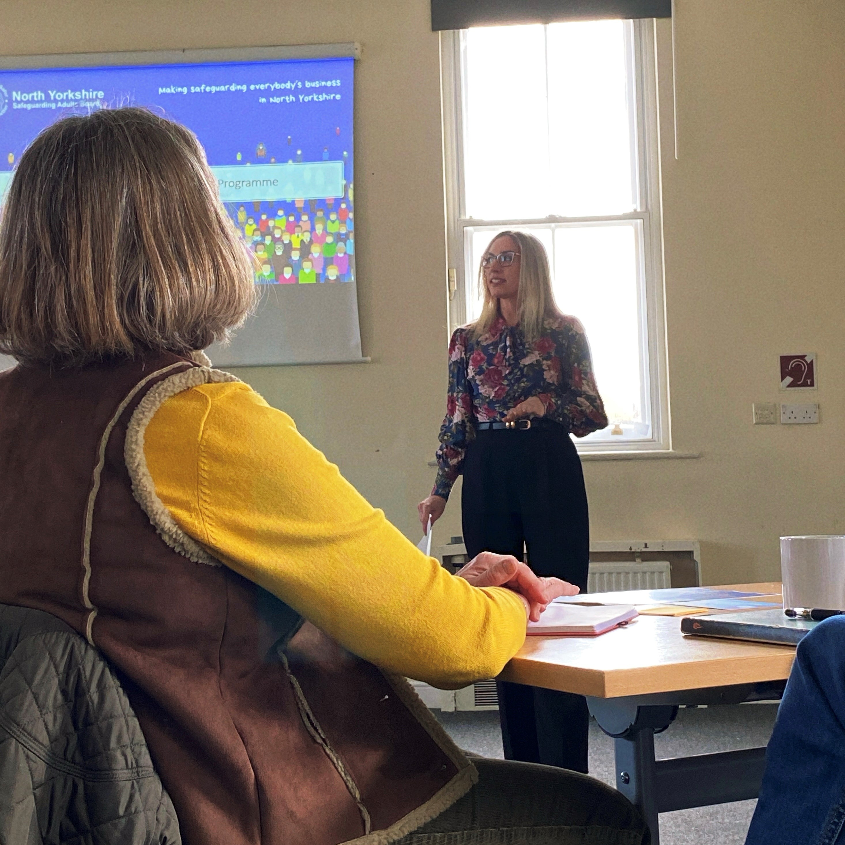 A blonde woman stands at the front of a room delivering a presentation on a screen about the workforce champions programme, as a toom of people watch on. 